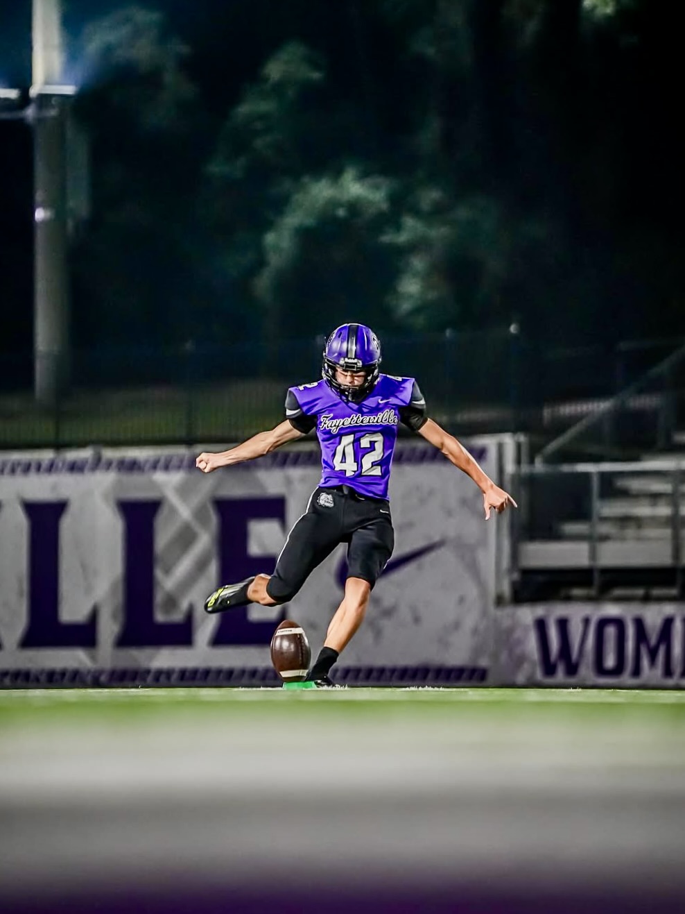 Ethan Farthing kicking off at night for Fayetteville High School in purple uniform.
